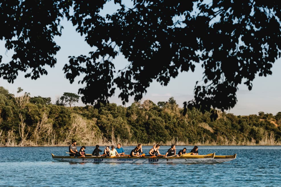 A long canoe full of paddlers on a calm lake under dark tree branches